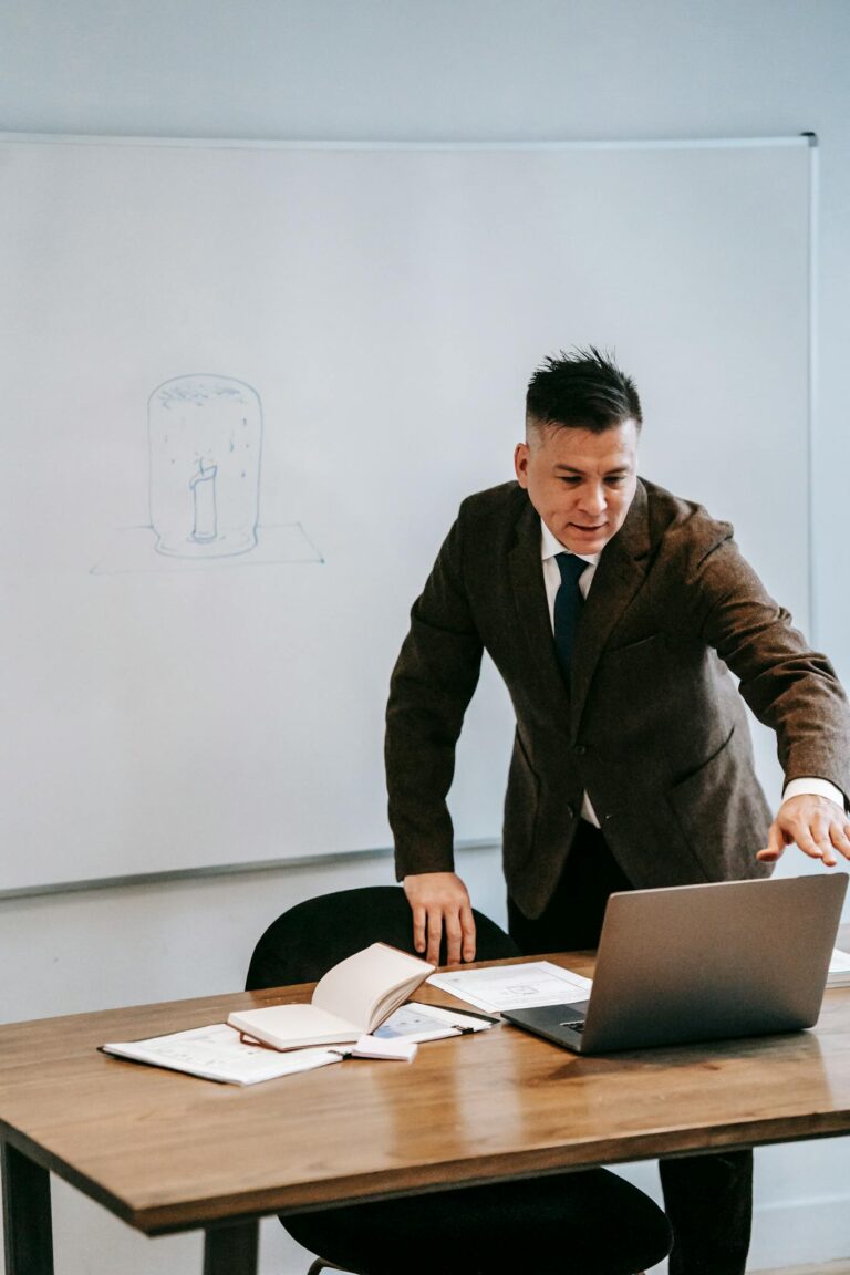 A professional man in a suit teaching online from an office setting with a laptop.